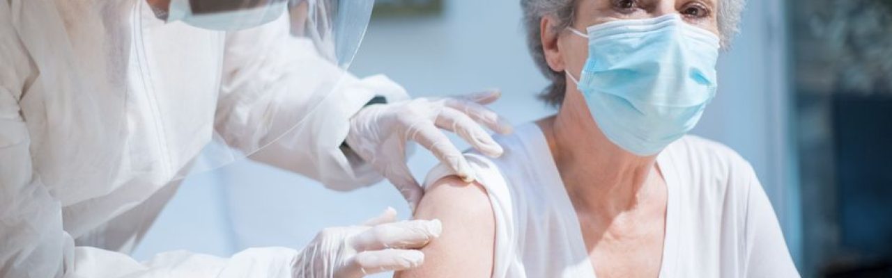 Senior woman wearing a mask getting vaccinated by a healthcare worker in PPE.